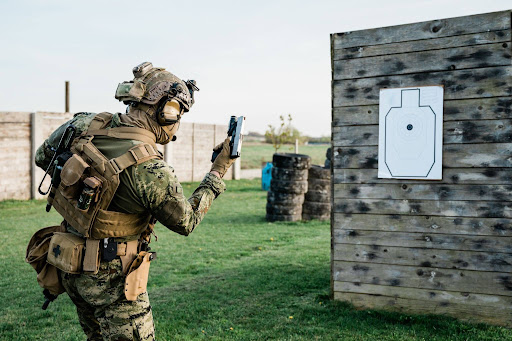 A person in full tactical gear is seen from behind, wearing a tan plate carrier layered over camouflage fatigues.